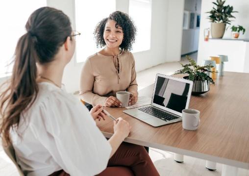 Two women sitting at a table in an office with coffe cups and an open laptop